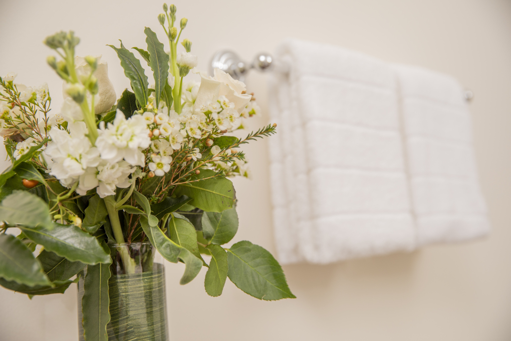 A vase with white flowers and green leaves on a table in front of a bathroom wall with white towels.